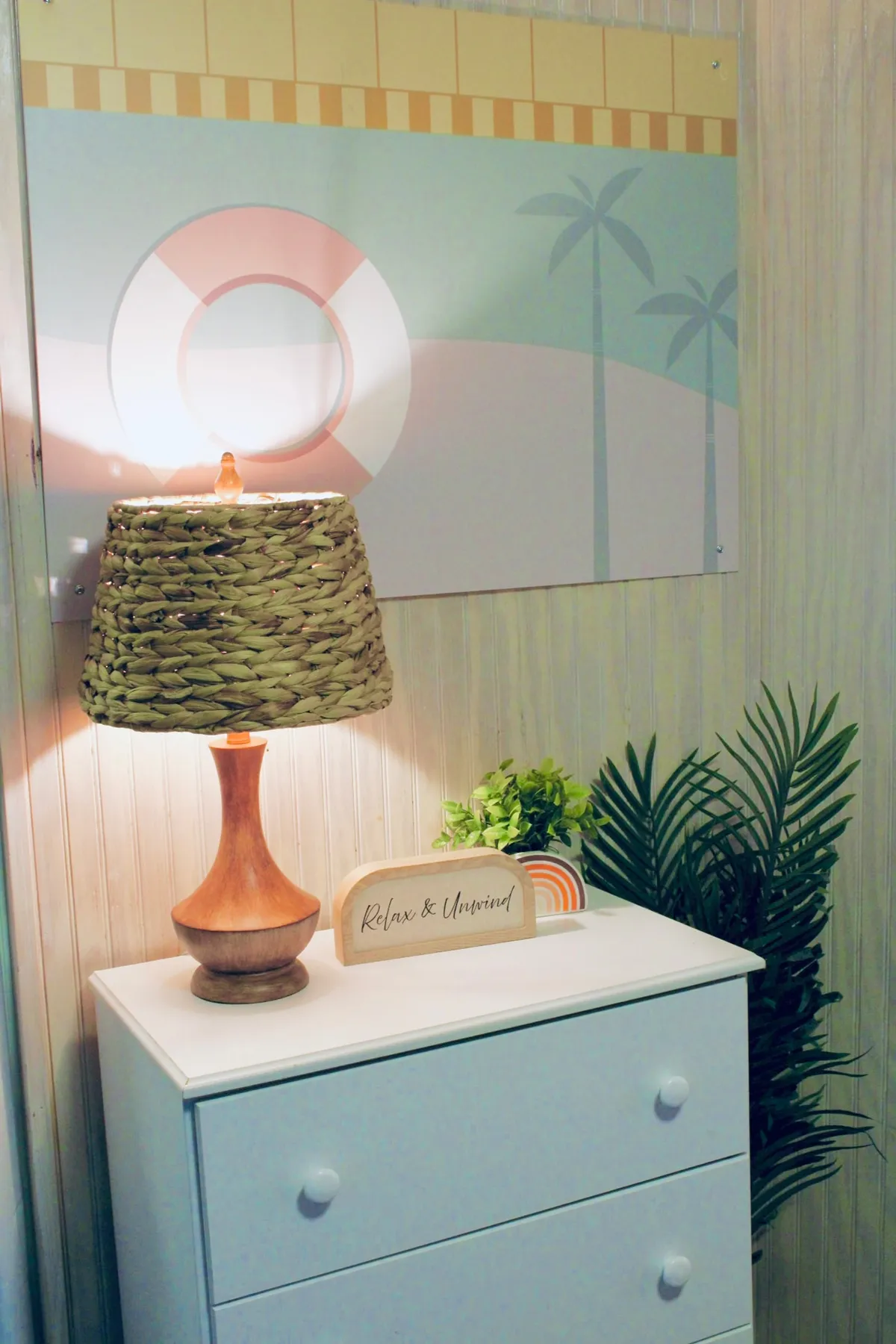A cozy corner with a table lamp, decorative sign, wall art, and a small plant, evoking a calming ambiance ideal for relaxation or preparing for the day ahead. The neutral color palette and soft lighting create a sense of comfort and retreat.