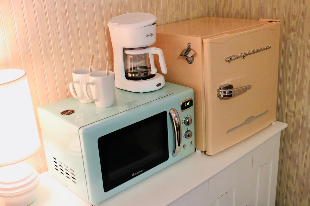 This image shows a bright, inviting kitchenette featuring a mint green microwave, a cream-colored coffee maker, and a retro pink refrigerator. White ceramic mugs are ready for use, and a soft lamp bathes the workspace in cozy light. The setup is compact yet thoughtfully organized, with enough counter space for quick meal prep or coffee breaks.
