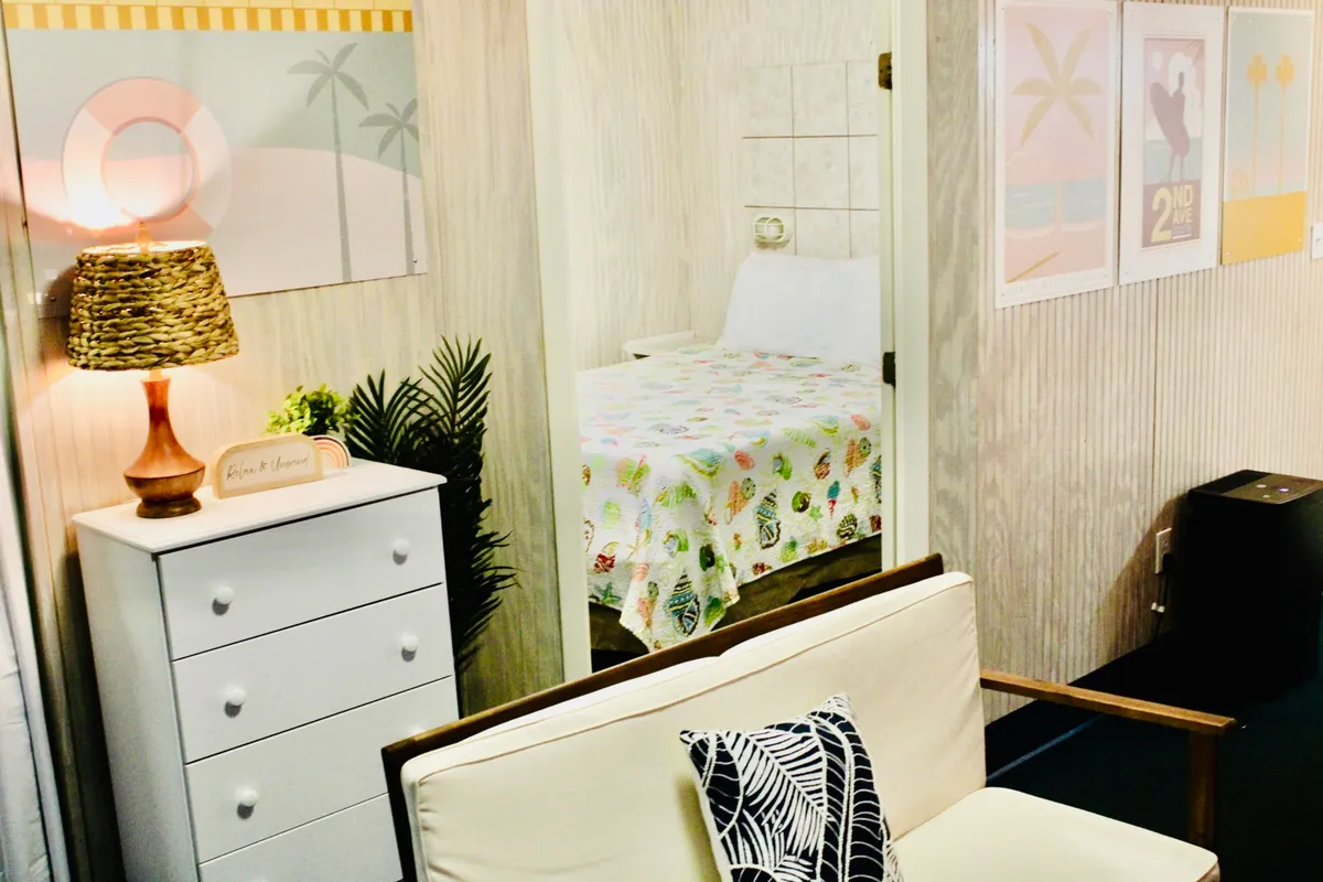 A sunny, cheerful bedroom with a woven lamp, colorful bedding, decorative plant, and playful art prints. The atmosphere is bright and welcoming, perfect for relaxing, reading, or socializing with friends or family—including four-legged guests.