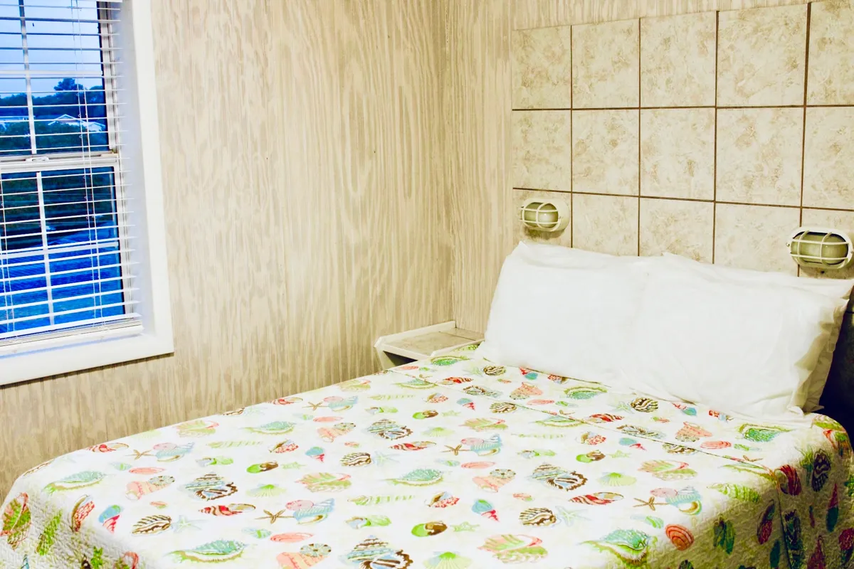 A cozy bedroom featuring a colorful quilt on the bed, soft window light filtering through blinds, and a nightstand with warm-lit lamps. The atmosphere is relaxing and homely, inviting restful moments after a day at the beach.